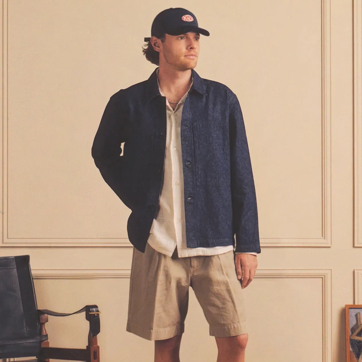 Man wearing a navy blue button-up jacket over a white shirt, beige shorts, and a black cap indoors.