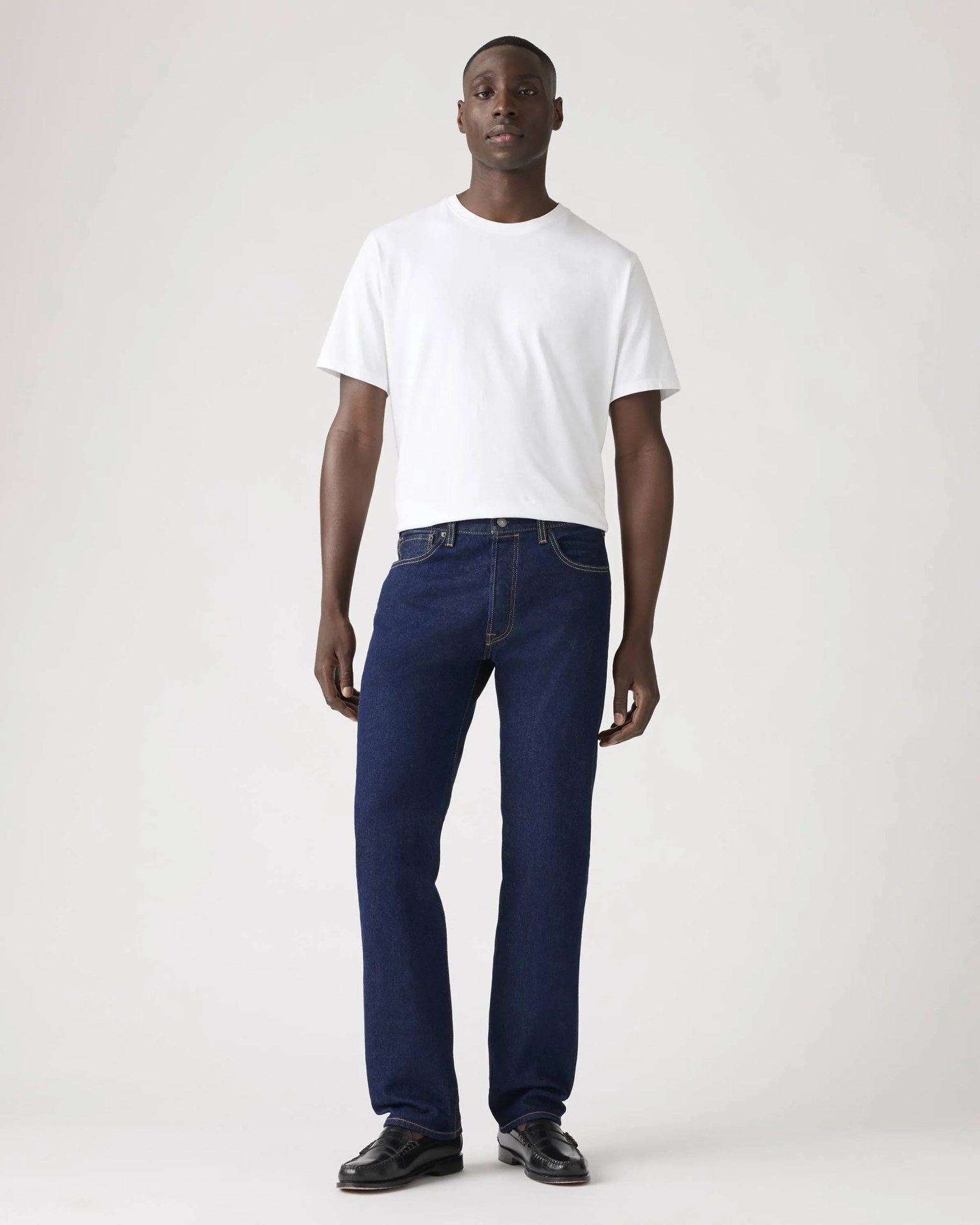 Man wearing a plain white short-sleeve t-shirt, dark blue jeans, and black leather loafers against a white background.