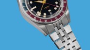 Close-up of a stainless steel wristwatch with a black dial, gold hands, red and blue GMT bezel, and a date window.