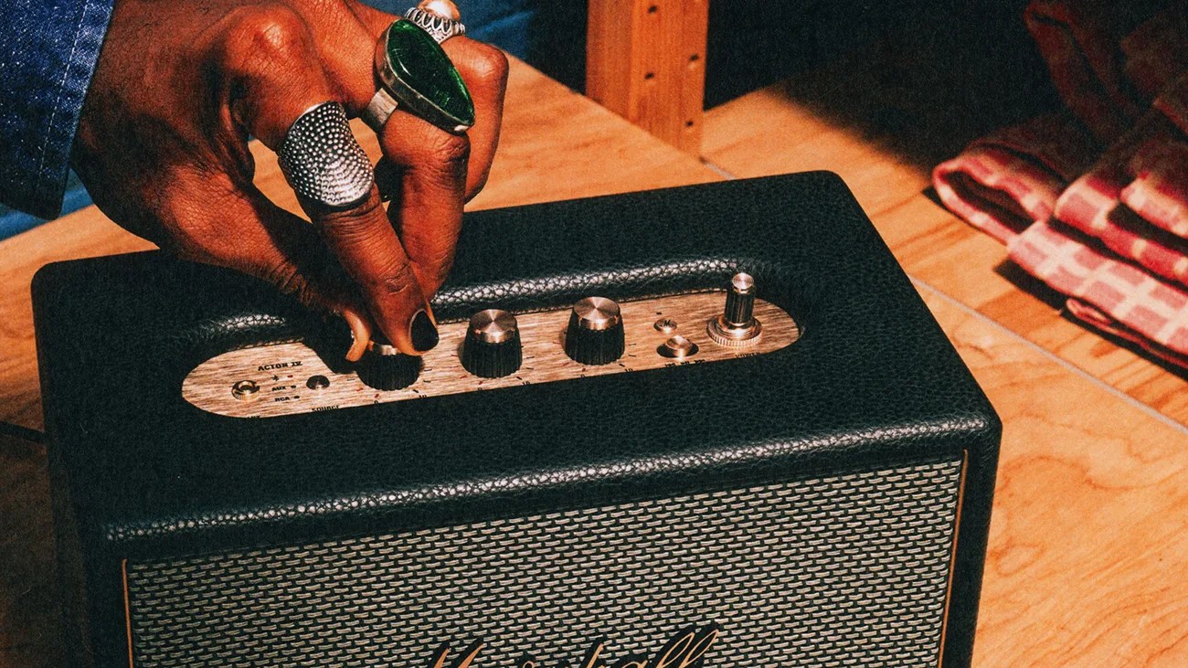 Hand with multiple rings adjusting a knob on a black Marshall speaker with wooden control panel.