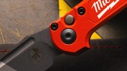 Close-up of a black folding knife blade with a logo and a red handle with visible screws.