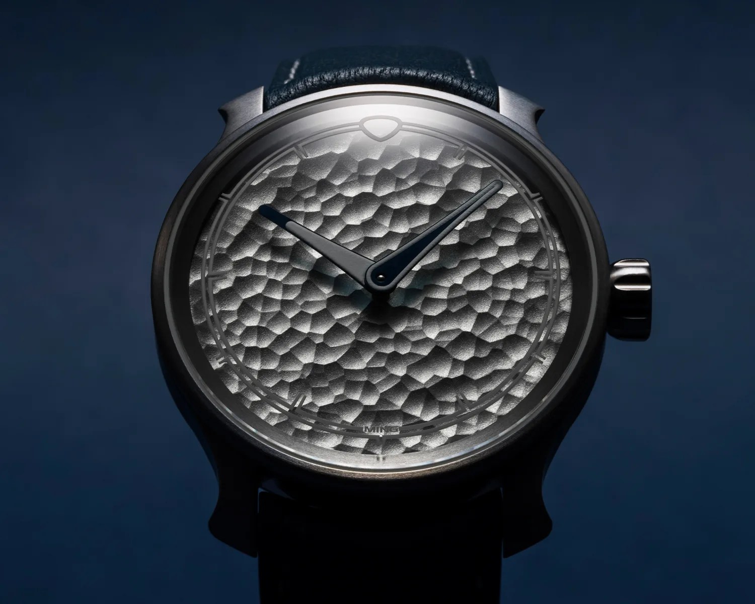 Round wristwatch with a textured silver dial, black hour and minute hands, and a black leather strap.