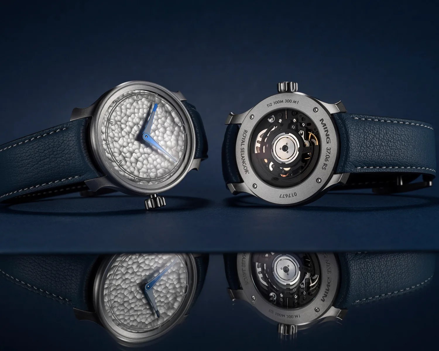 Two titanium wristwatches with textured silver dials, blue hands, and blue leather straps on a reflective dark surface.