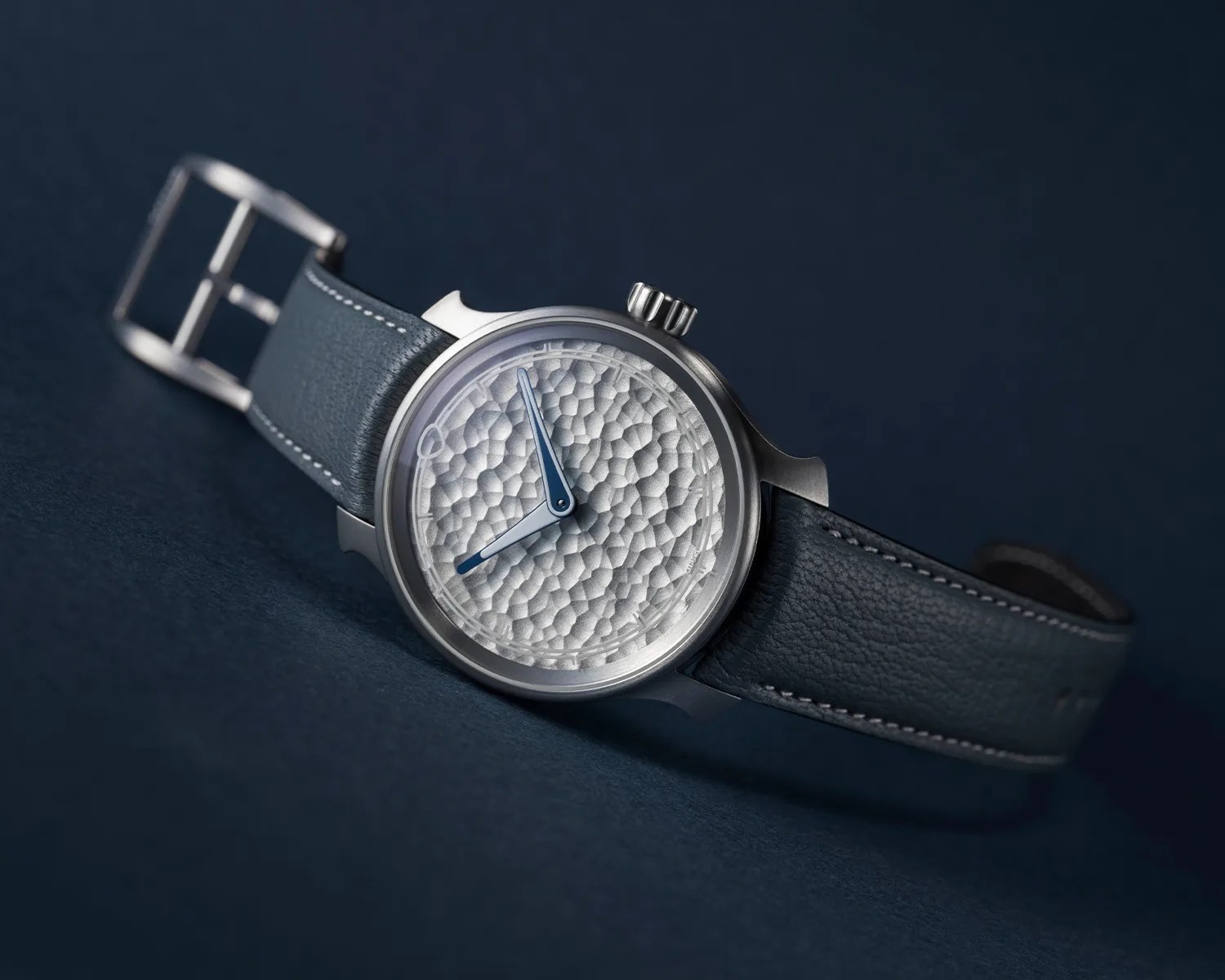 Silver wristwatch with textured dial, blue hands, and dark blue leather strap on a dark blue surface.