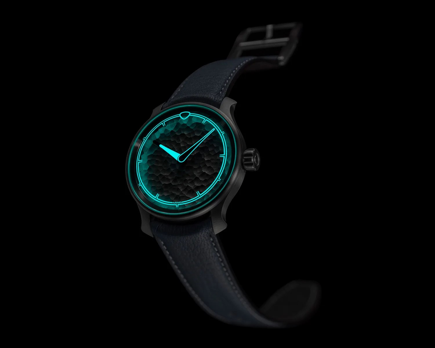 Black wristwatch with textured dial and glowing turquoise hour markers and hands on a dark leather strap.