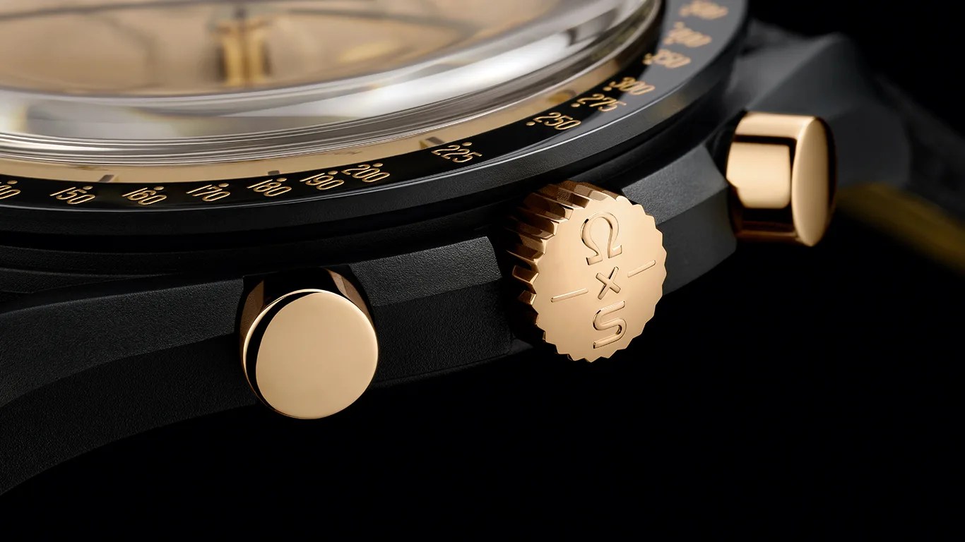Close-up of a black watch case with gold crown engraved with "Ω x S" and two gold pushers.