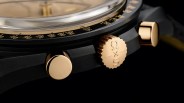 Close-up of a black watch case with gold crown engraved with "Ω x S" and two gold pushers.