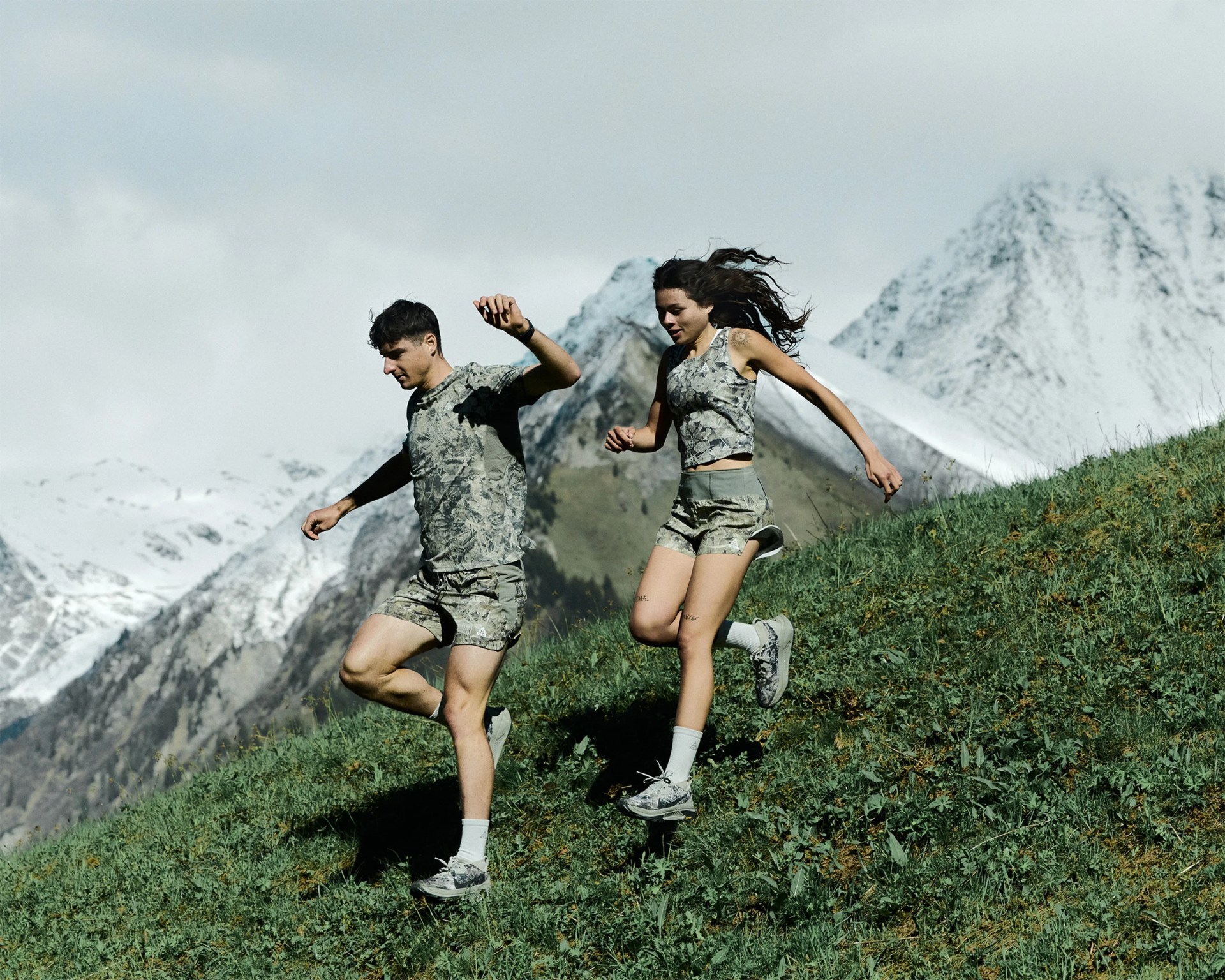Two people in matching gray floral activewear running downhill on a grassy slope with snow-capped mountains in the background.