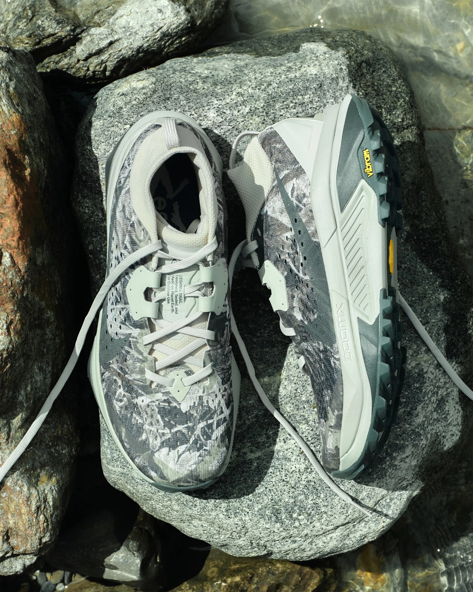 Pair of gray and white trail running shoes with Vibram soles on a large rock near water.
