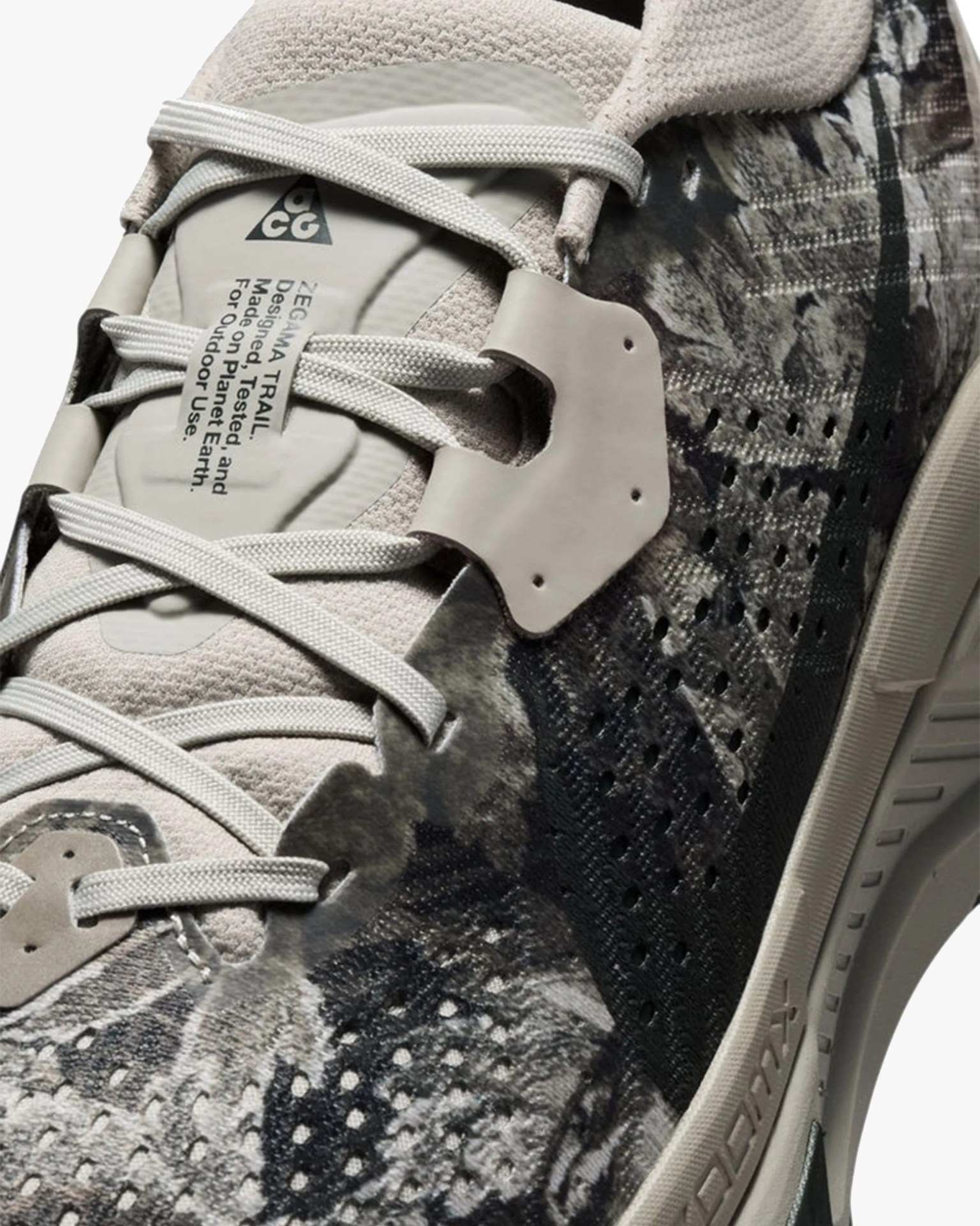 Close-up of a beige and black patterned sneaker with white laces and a label reading "ZEGAMA TRAIL Designed, Tested, and Made on Planet Earth For Outdoor Use.