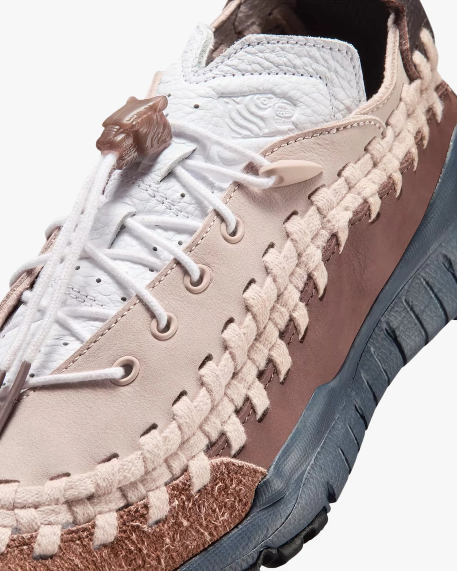 Close-up of a beige and brown sneaker with white laces, a tiger head lace lock, and a blue textured sole.