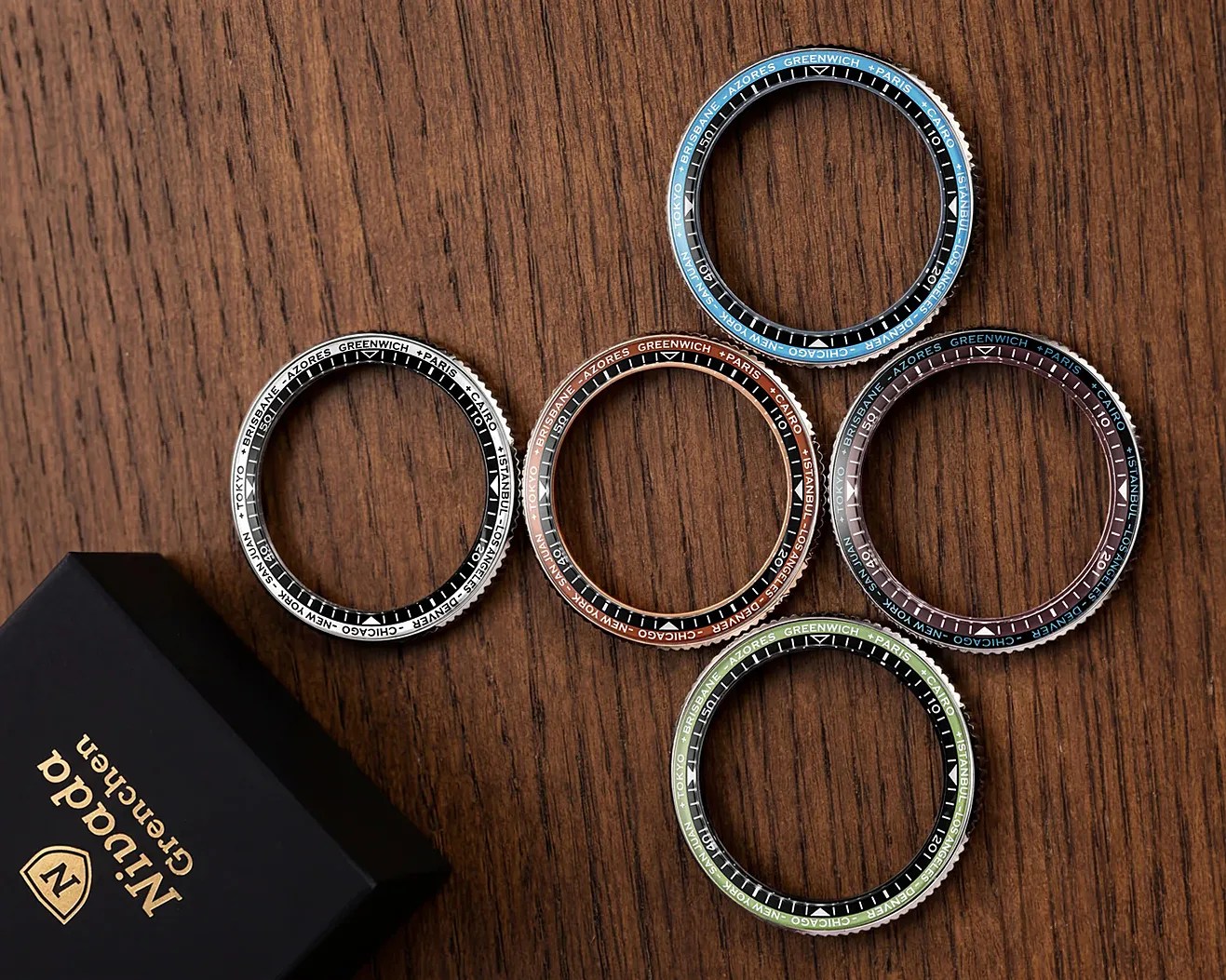 Five colorful watch bezels with city names and numbers on a wooden surface next to a black box with gold "Nivada Grenchen" branding.