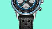Close-up of a wristwatch with a black and blue bezel, black dial with three subdials, and a black perforated leather strap.