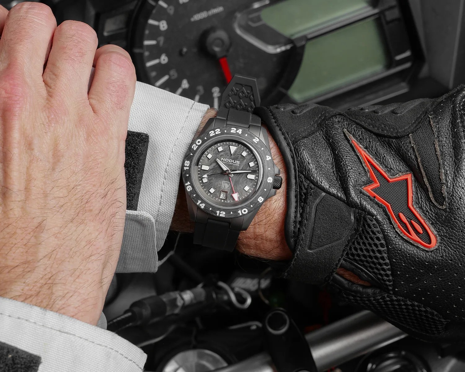Black Nodus wristwatch with white hour markers and a red-tipped GMT hand worn on a wrist with a black glove.
