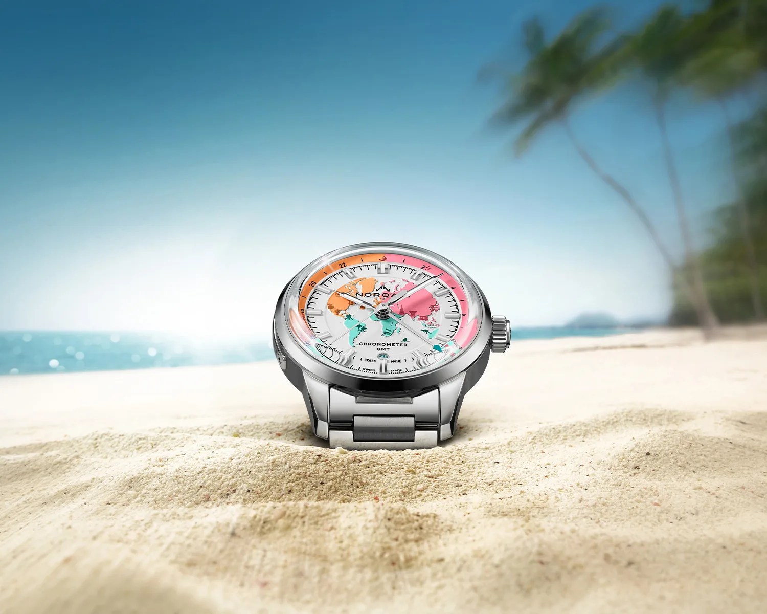 Silver metal wristwatch with a colorful world map dial resting on sandy beach with ocean and palm trees in background.