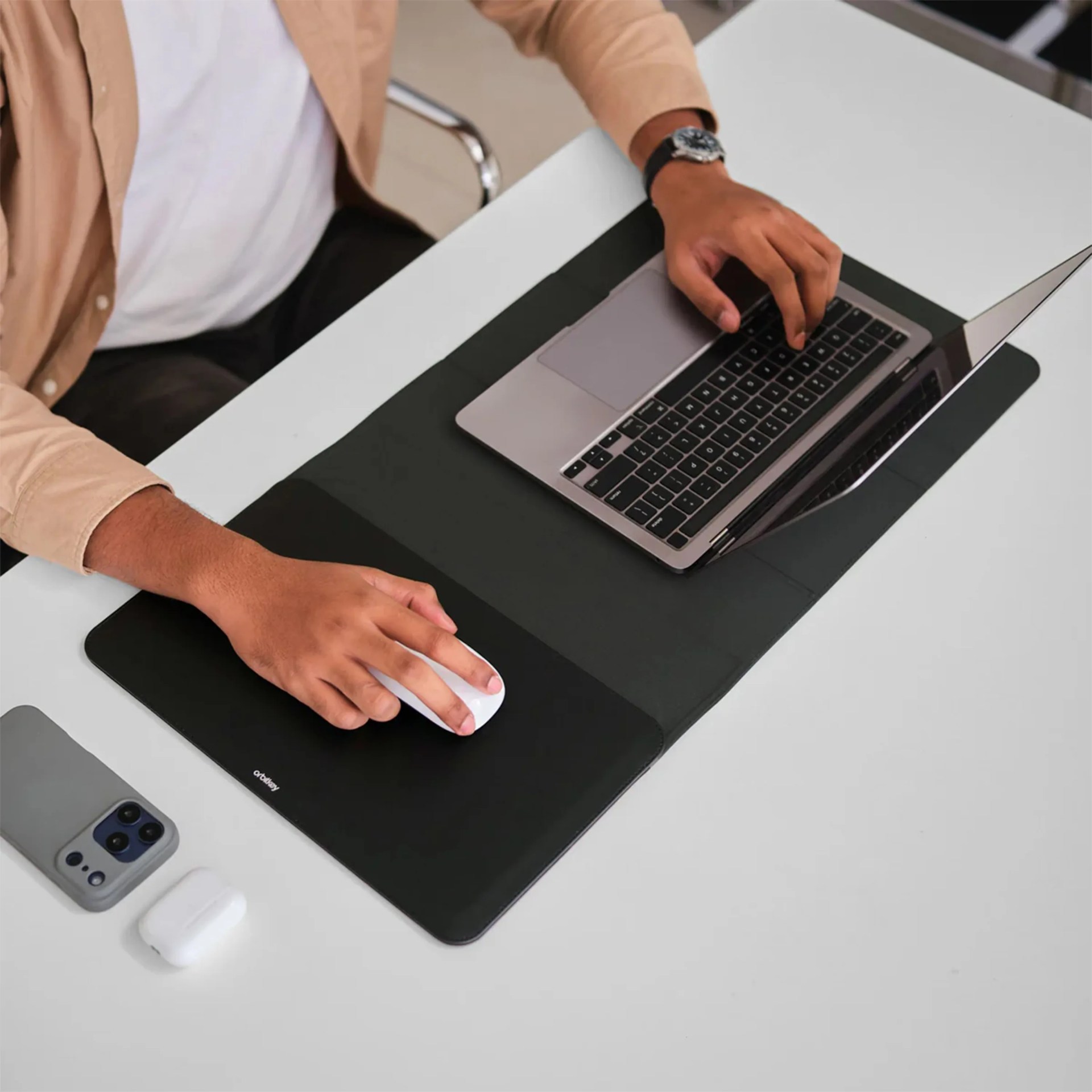 Person using a silver laptop on a black desk mat with a white wireless mouse and smartphone nearby.