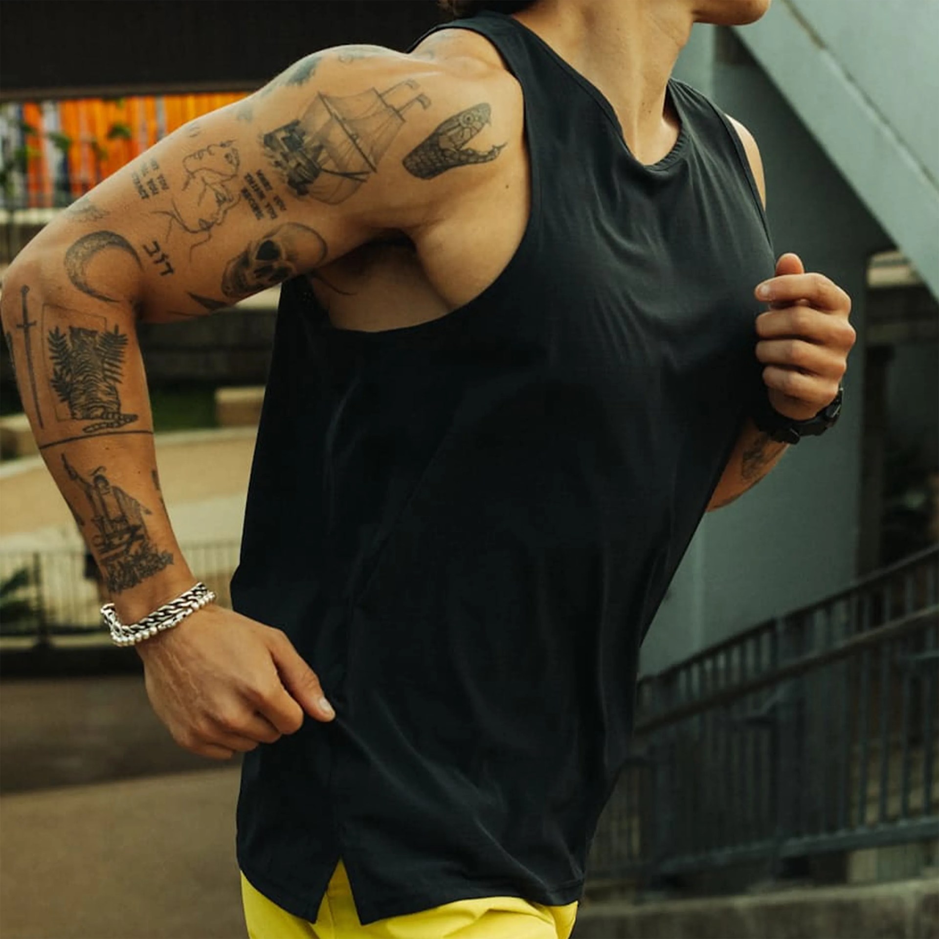 Close-up of a person running wearing a black sleeveless shirt, yellow shorts, and multiple tattoos on their arm.