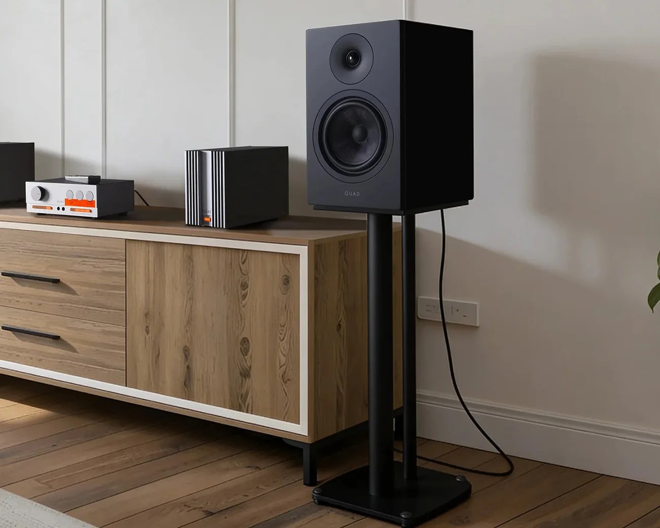 Black bookshelf speaker on a black metal stand next to a wooden cabinet with audio equipment on top.