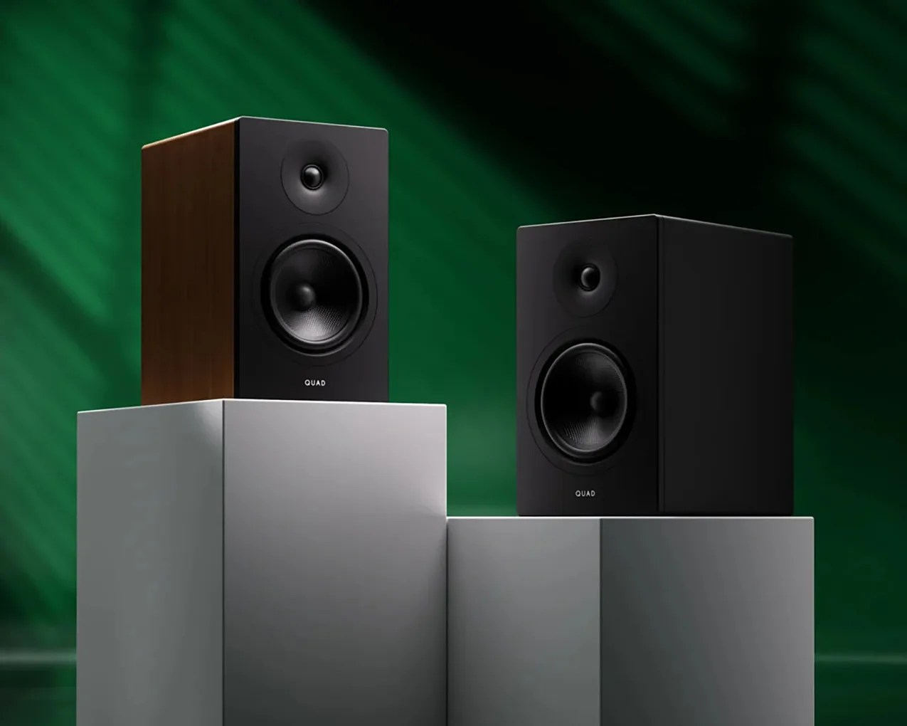 Two black Quad bookshelf speakers, one with wood finish sides, displayed on gray pedestals against a green background.