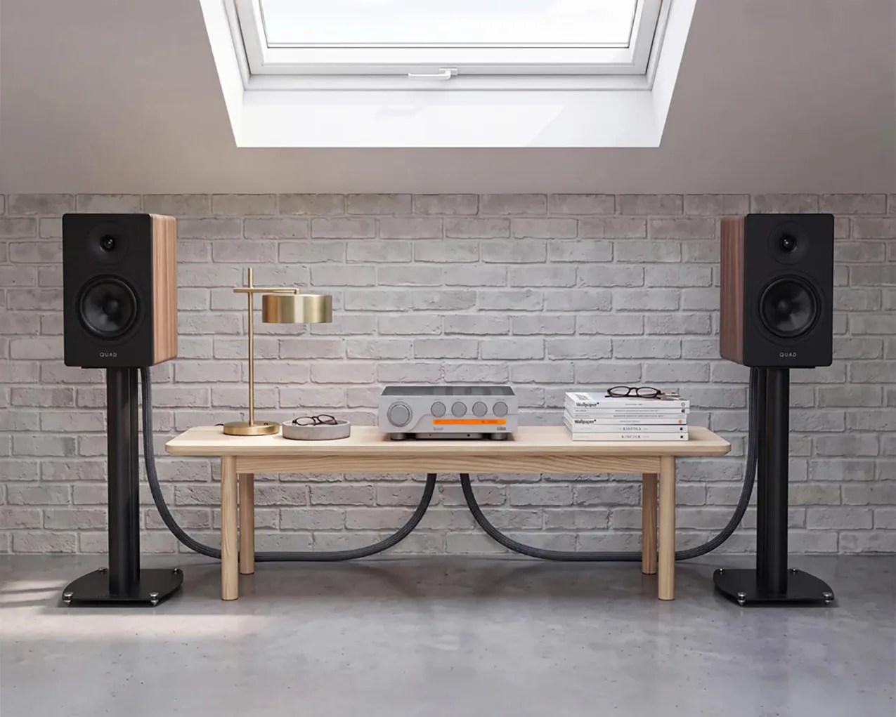 Pair of black and wood Quad speakers on black stands flanking a light wood bench with a silver amplifier, gold lamp, and stacked books.
