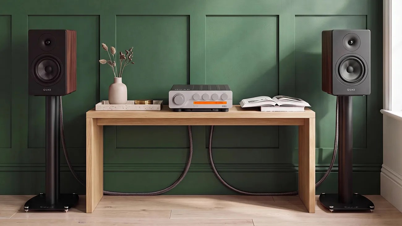 Pair of black and wood Quad speakers on black stands flanking a wooden table with a Quad amplifier, vase with dried flowers, and open book.