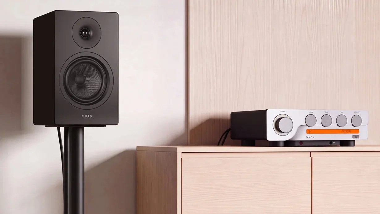 Black Quad bookshelf speaker on a stand next to a black and silver Quad amplifier on a wooden cabinet.