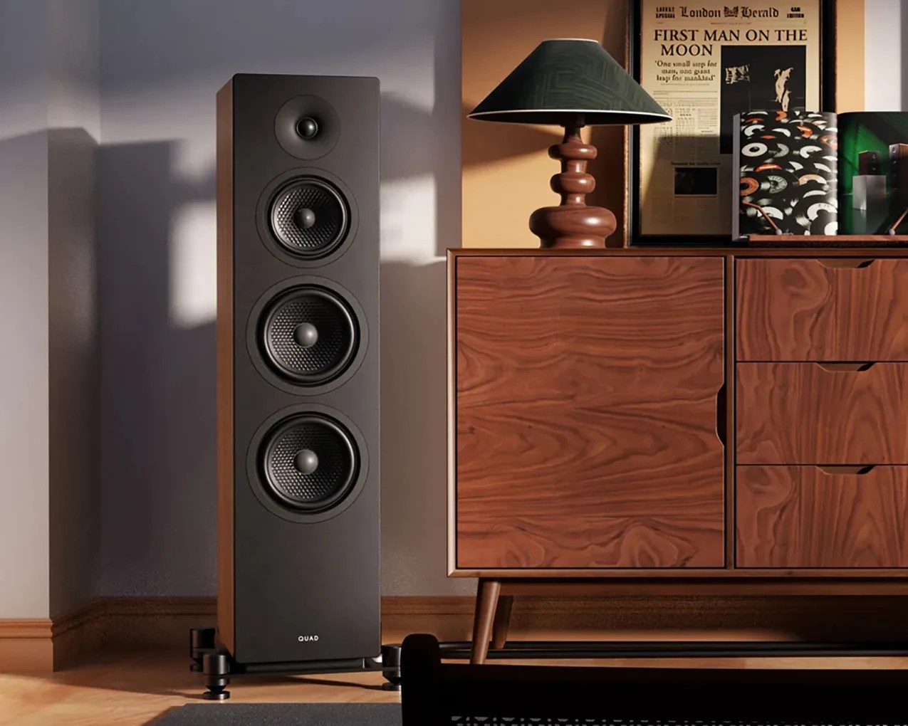 Tall black floor-standing speaker with four drivers next to a wooden sideboard with a lamp and framed newspaper.