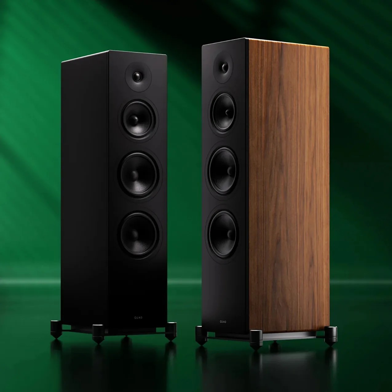 Pair of tall floor-standing speakers with black front panels and one with wood veneer side panel on a green background.
