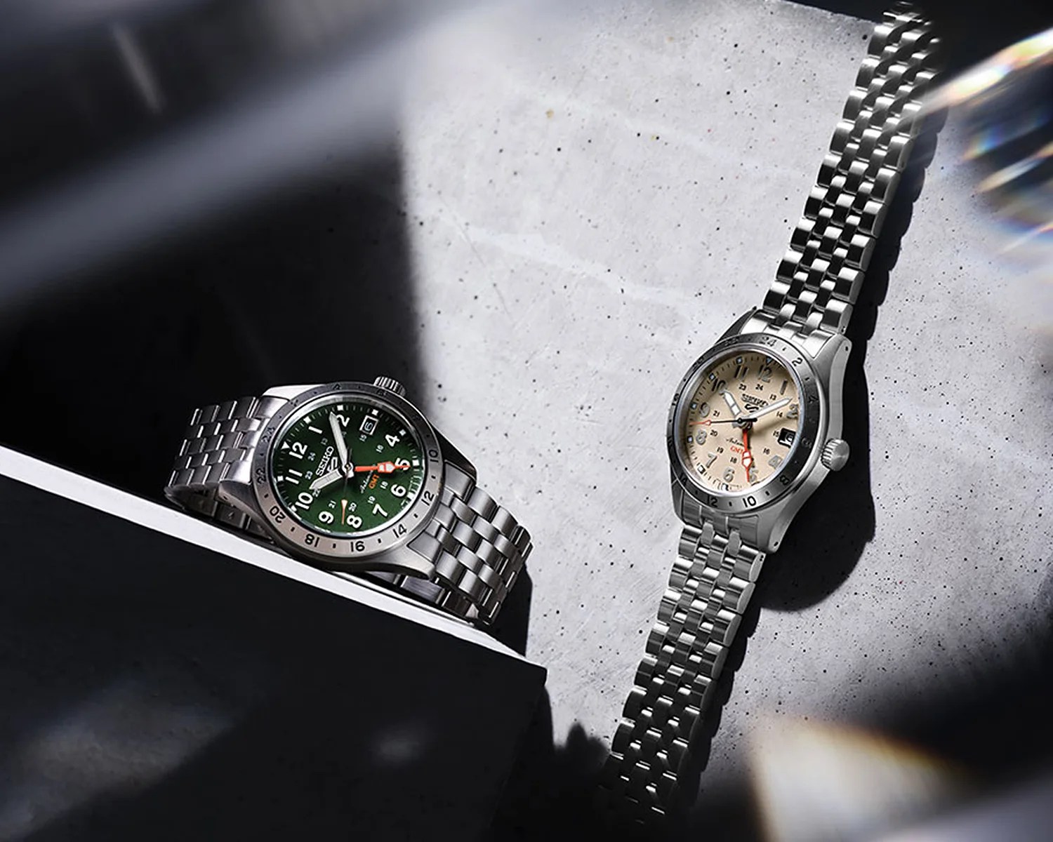 Two Seiko wristwatches with stainless steel bracelets, one with a green dial and the other with a beige dial, on a concrete surface.