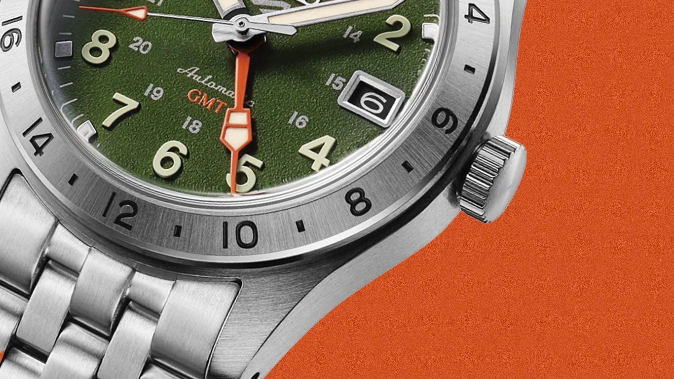 Close-up of a stainless steel watch with a green textured dial, orange GMT hand, and date window at 3 o'clock.