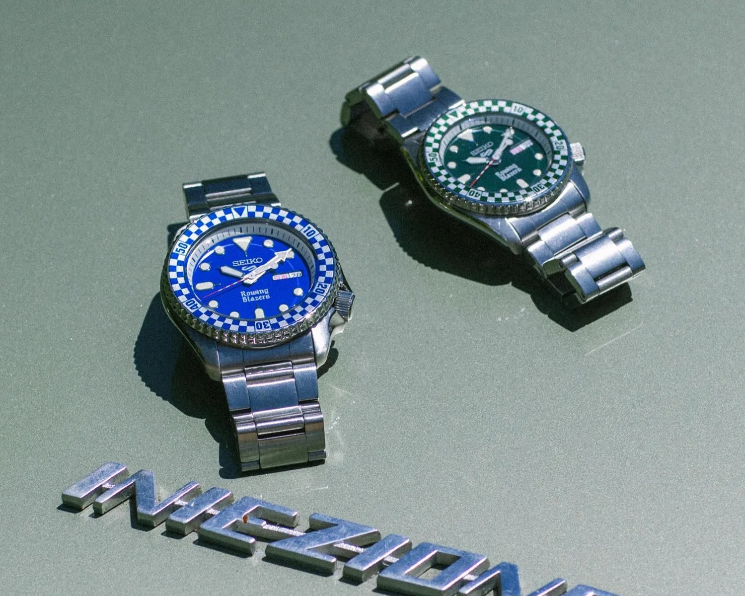 Two Seiko wristwatches with checkered bezels, one blue and one green, on a green car