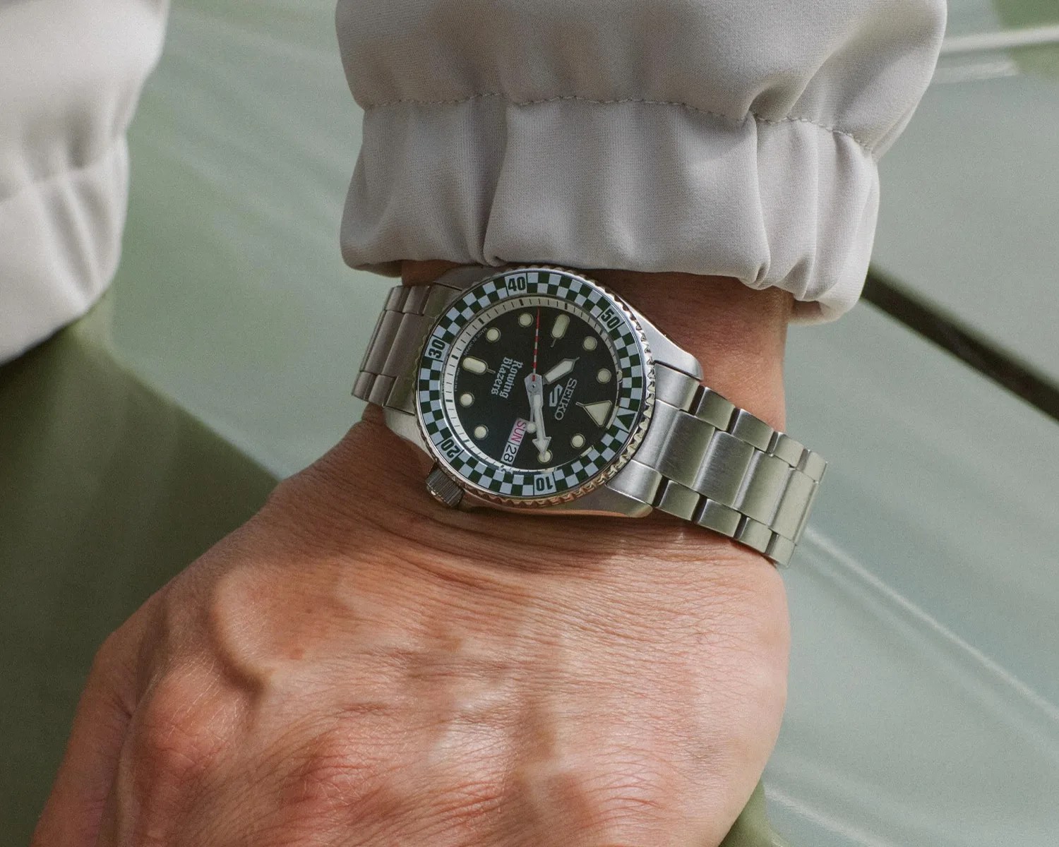 Silver Seiko wristwatch with black dial and green-and-white checkered bezel on a wrist.