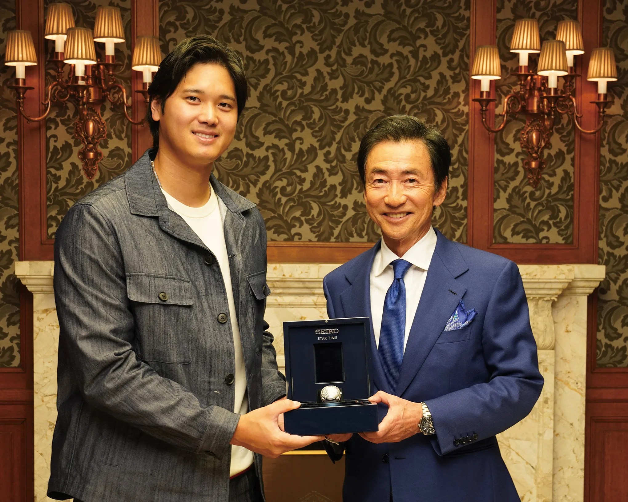 Two men smiling and holding a Seiko Star Time watch box in a room with ornate wallpaper and wall sconces.