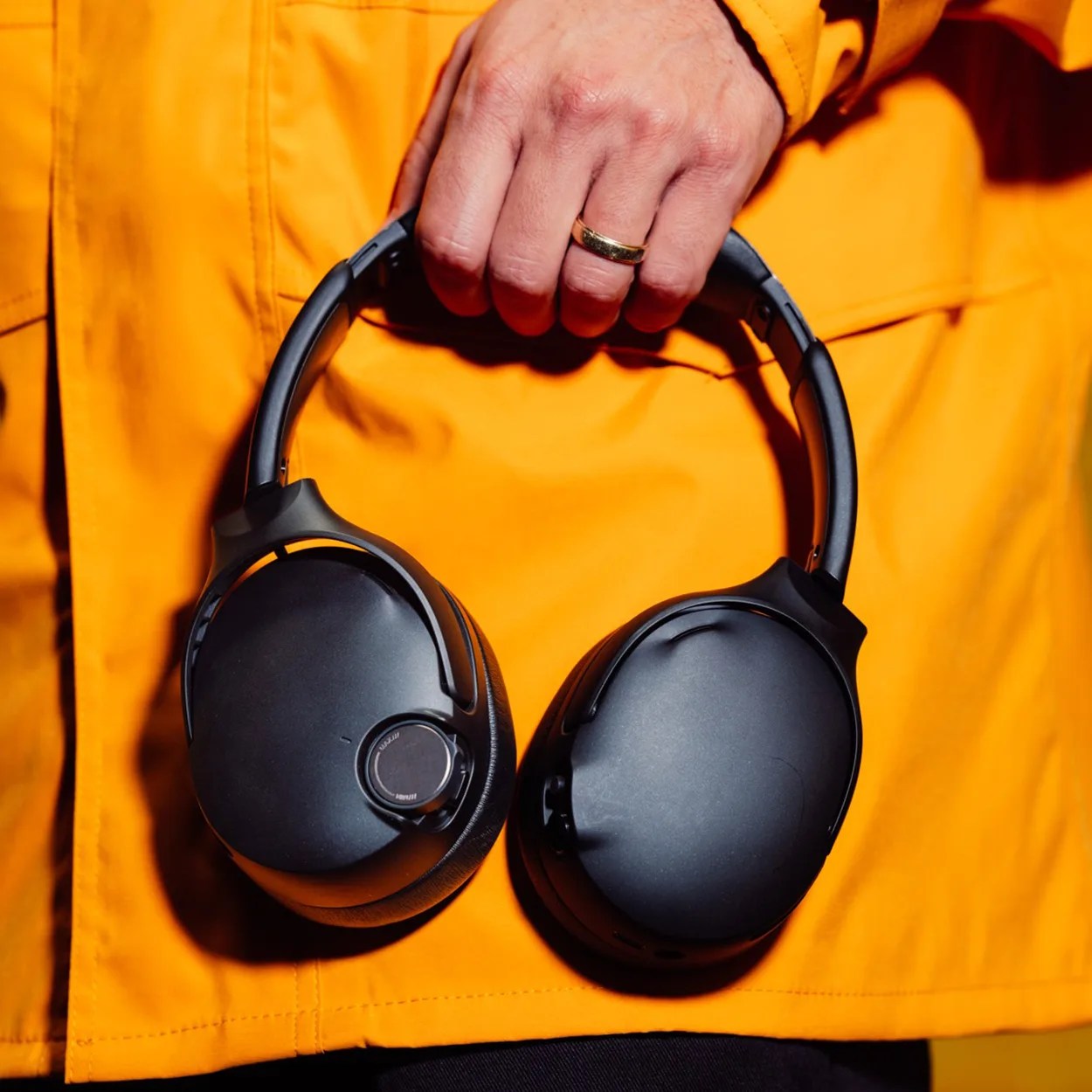 Black over-ear headphones held in a hand wearing a gold ring and an orange jacket.