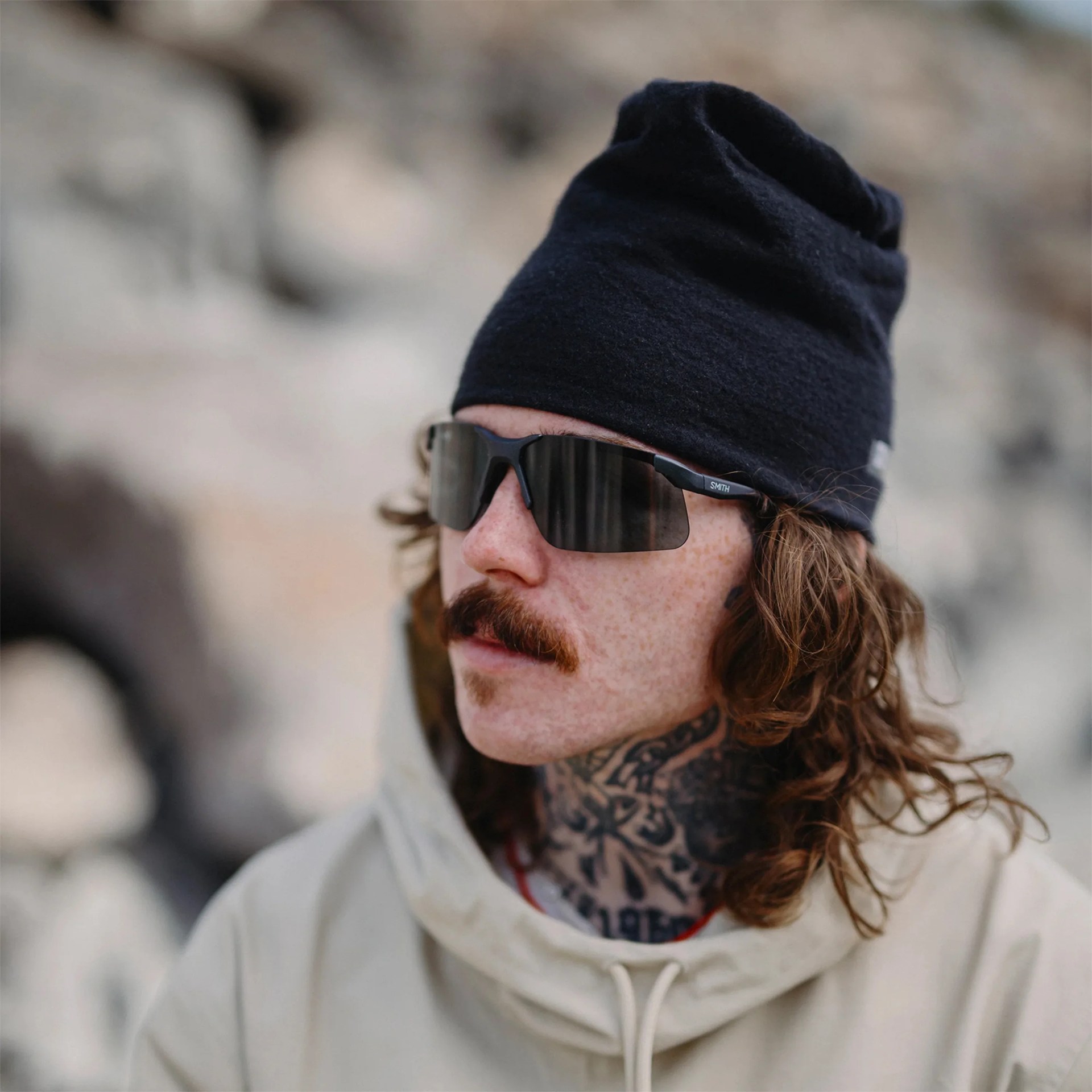 Close-up of a person wearing a black beanie, black wraparound sunglasses, and a light-colored hoodie with visible neck tattoos.