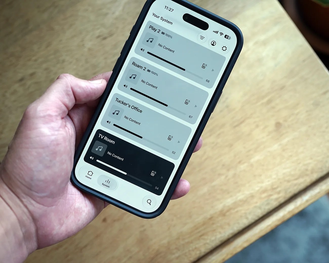Hand holding a smartphone displaying a home audio system app with multiple rooms listed and no content playing.
