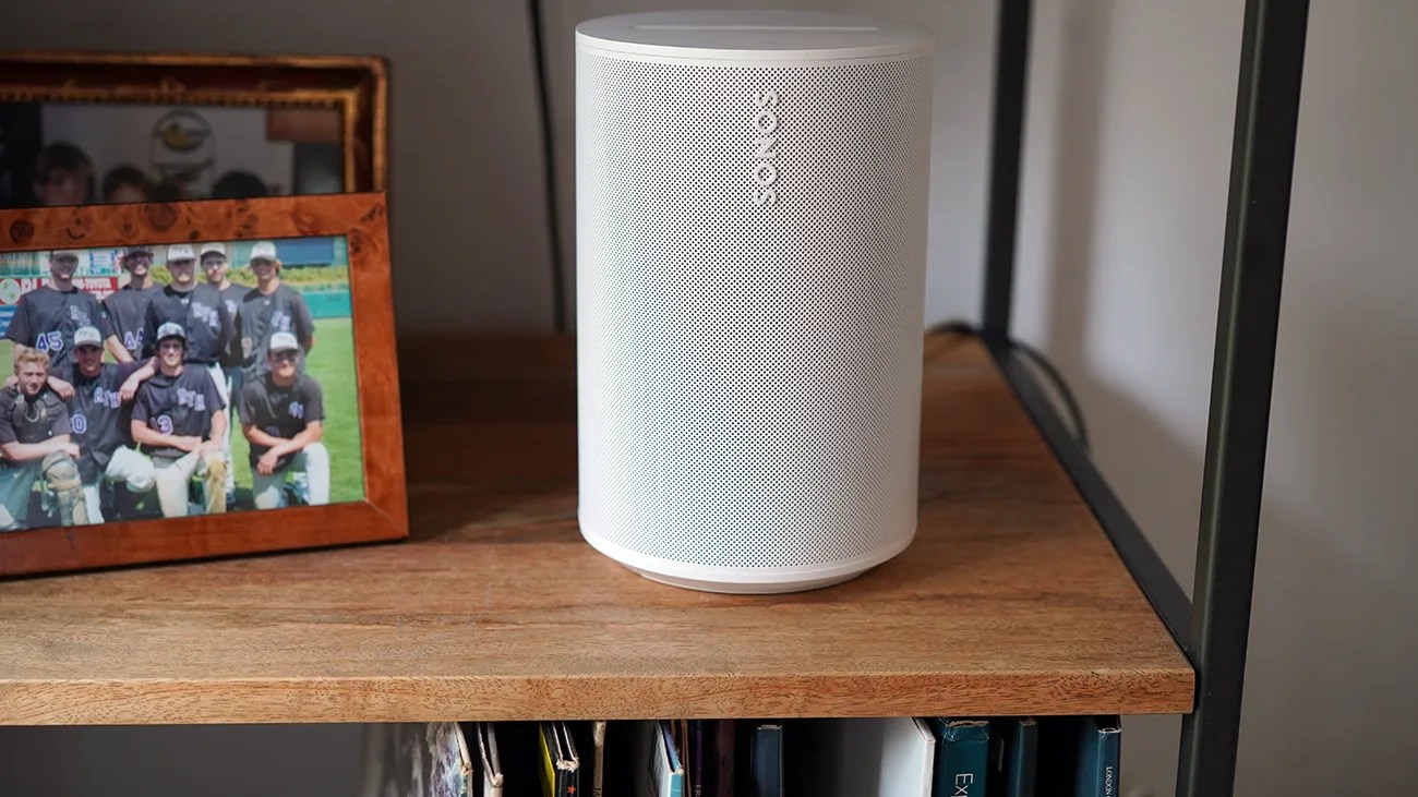 White Sonos wireless speaker on a wooden shelf next to a framed photo.