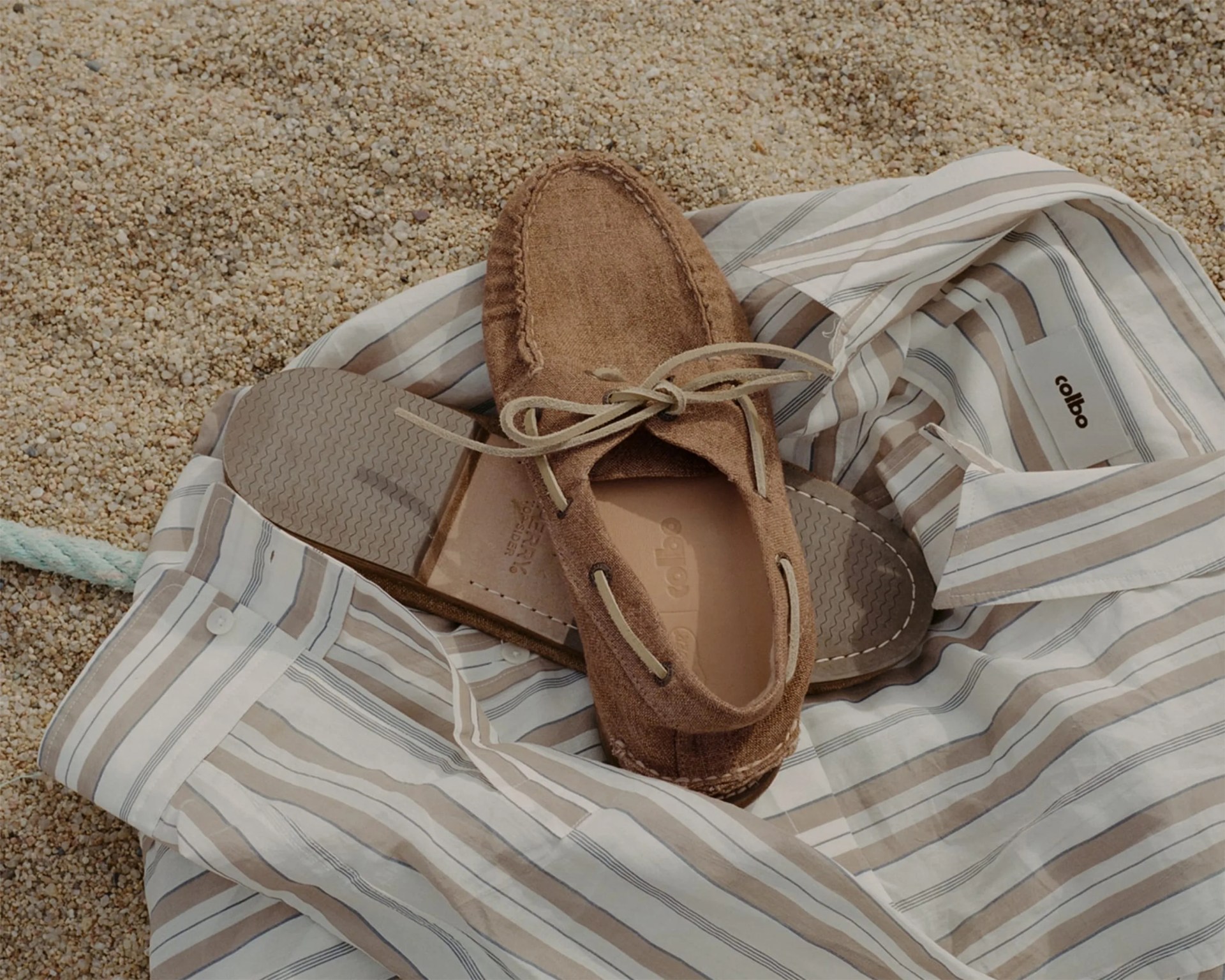 Brown suede moccasin shoe with beige laces on a striped beige and white shirt on gravel.