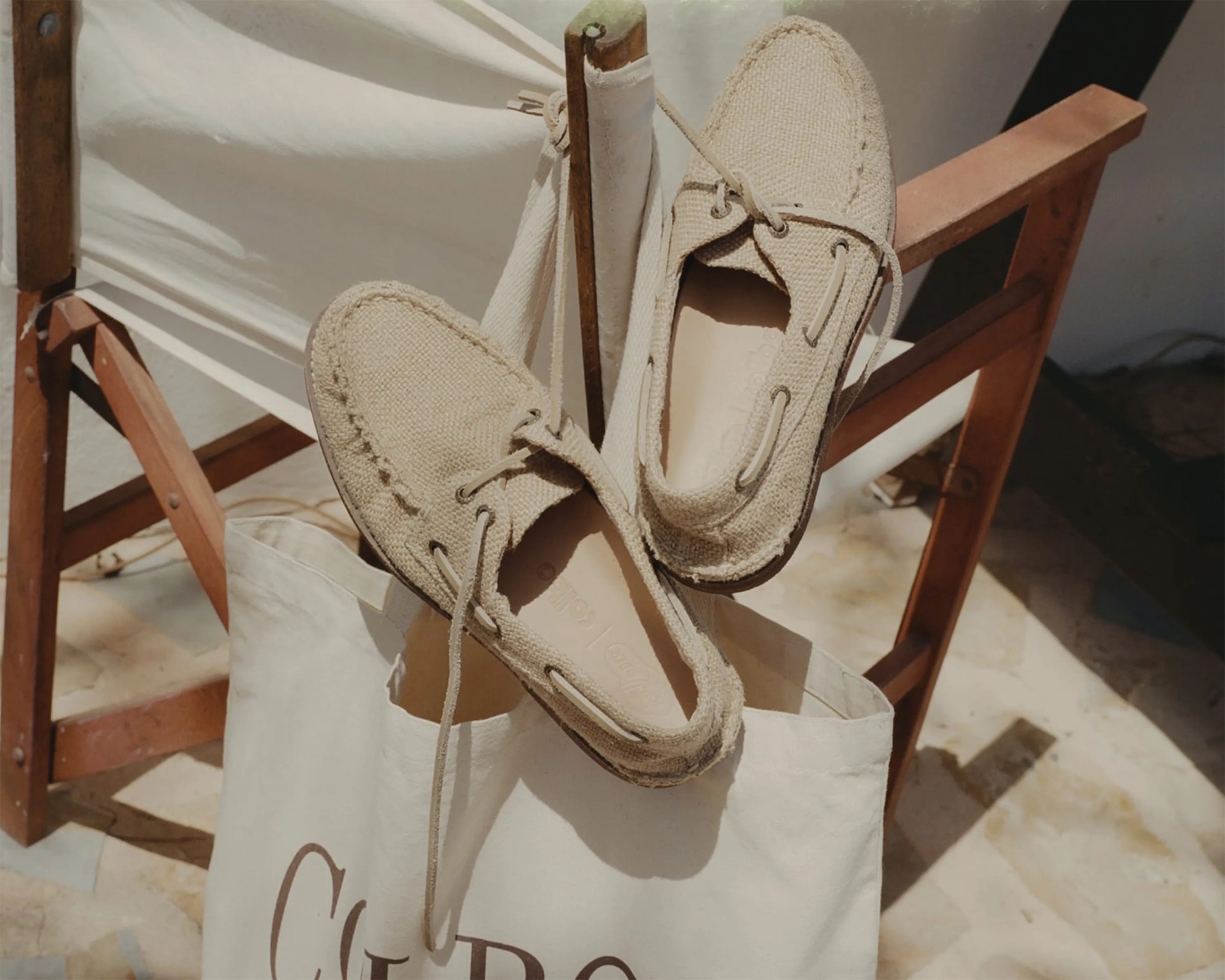 Beige woven moccasin loafers with laces resting on a white canvas tote bag on a wooden chair.