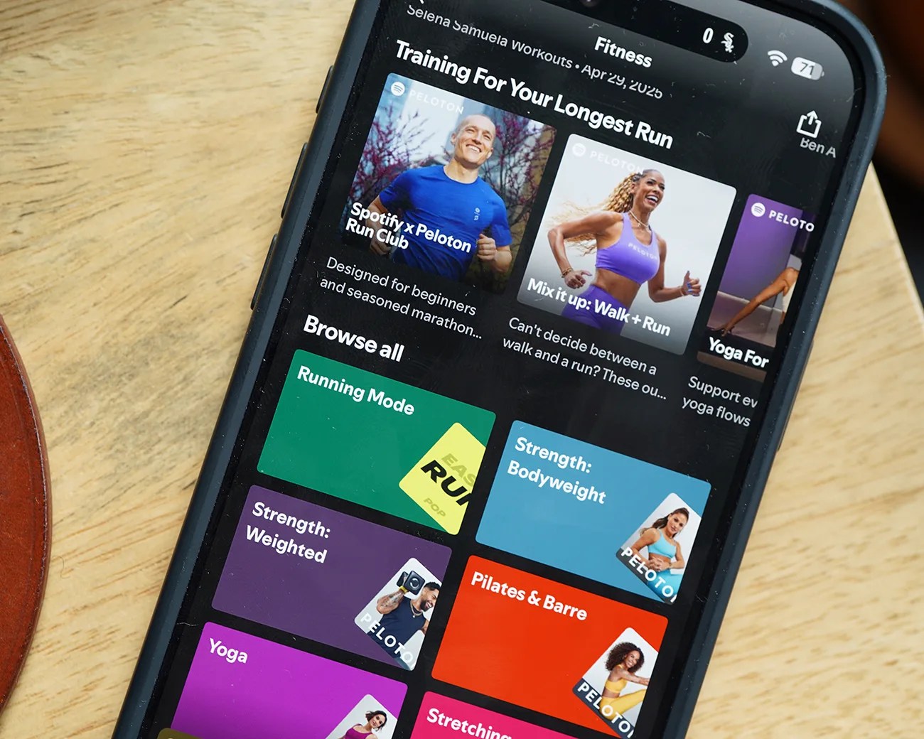Smartphone screen showing a fitness app with workout categories like Running Mode, Strength, Pilates, and Yoga on a wooden surface.