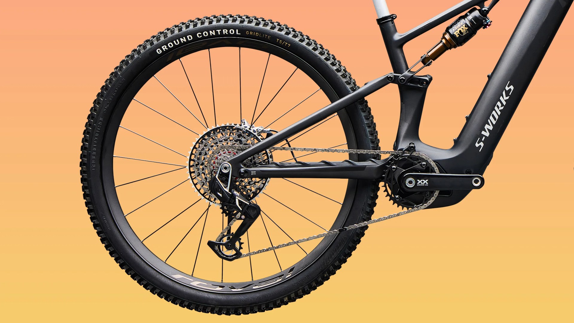 Close-up of a black S-Works mountain bike rear wheel, drivetrain, and suspension against an orange gradient background.
