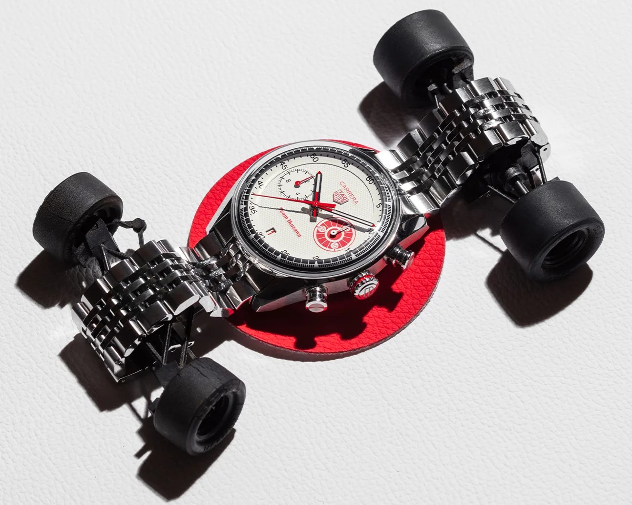 Silver TAG Heuer Carrera watch with white and red dial, attached to a miniature black race car chassis with rubber tires.