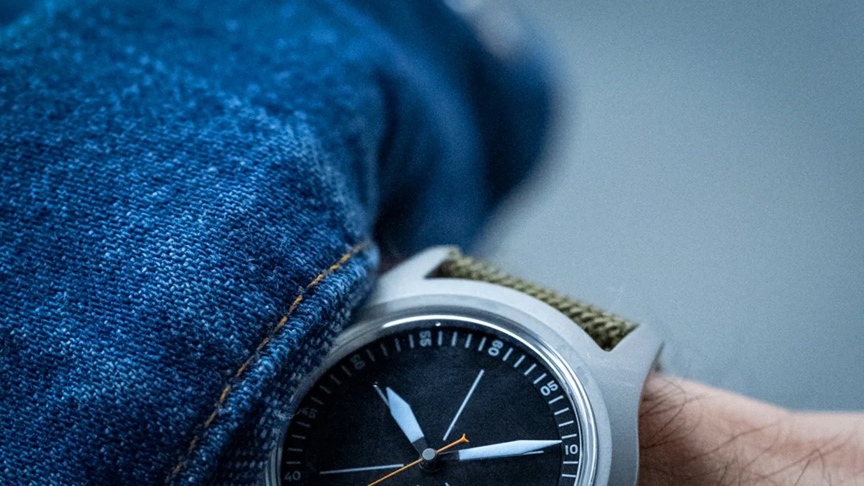 Close-up of a wristwatch with a black dial, white hands, and an olive green fabric strap worn under a blue denim sleeve.