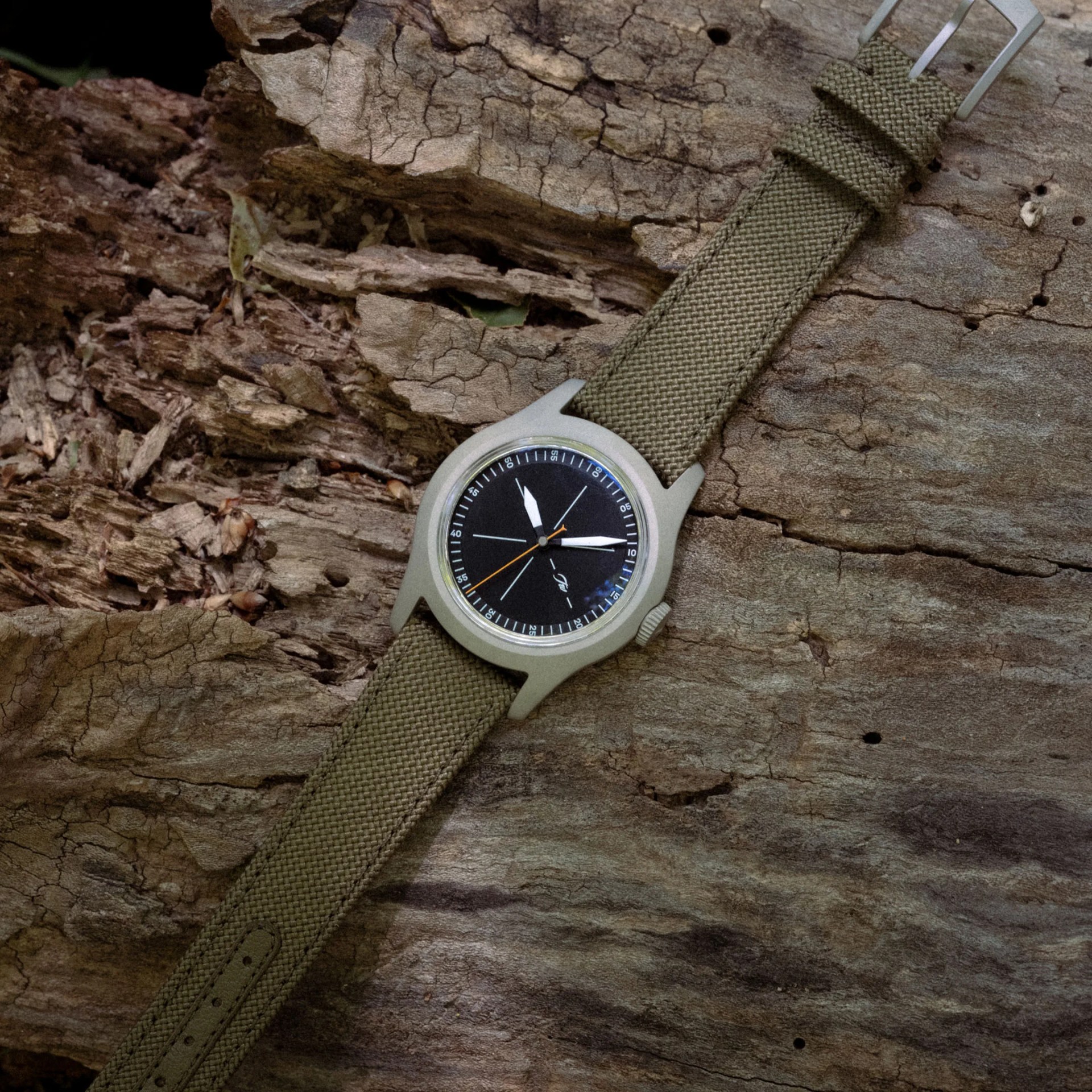 Watch with black dial, orange second hand, and olive green woven fabric strap on cracked wood surface.