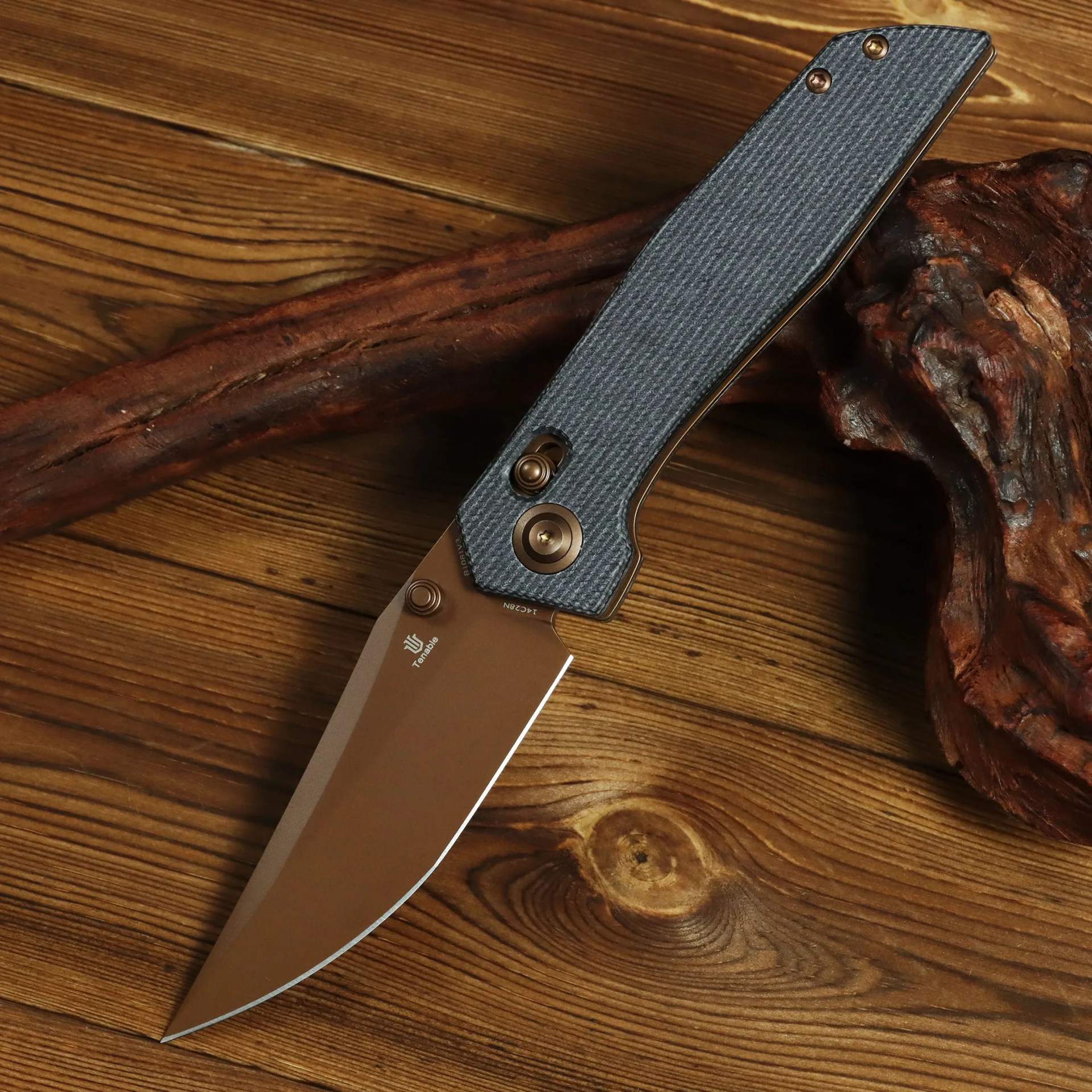 Folding knife with bronze blade and textured gray handle resting on wooden surface with a piece of driftwood.