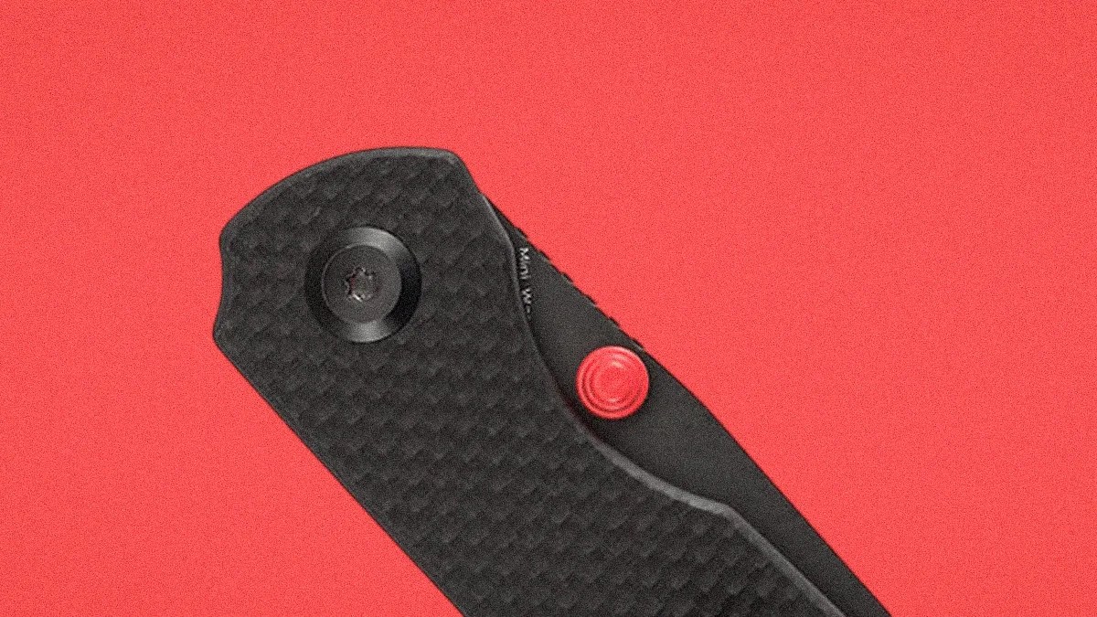 Close-up of a black textured folding knife handle with a red circular button on a red background.