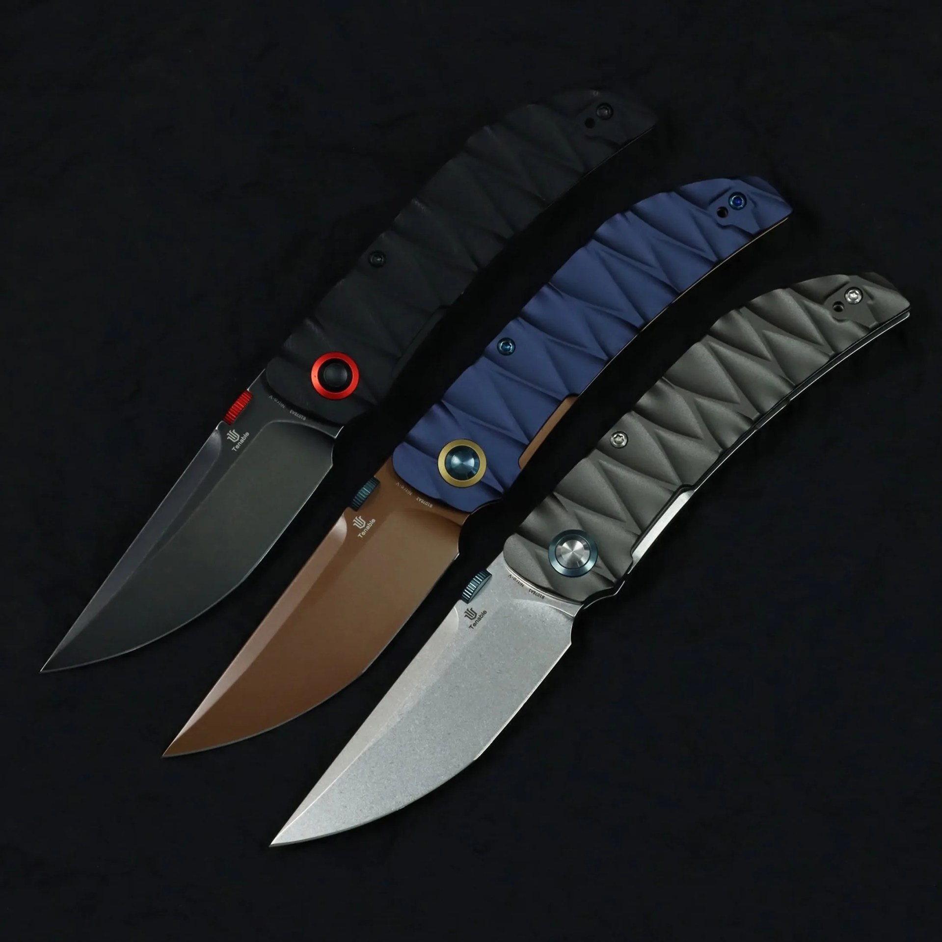 Three folding knives with textured handles in black, blue, and gray, each with a different colored pivot ring, laid on a black surface.
