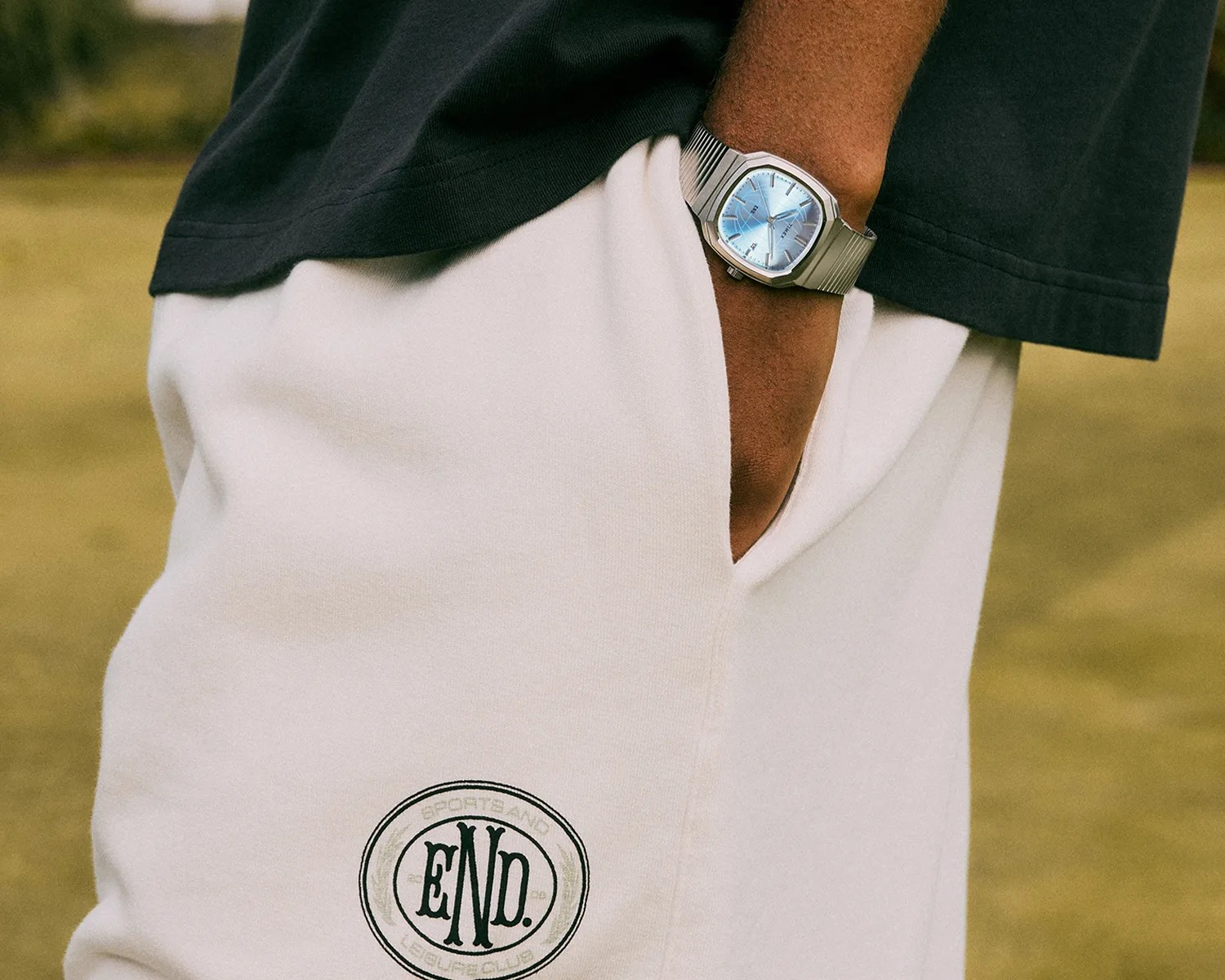 Silver wristwatch with a blue dial worn on a wrist, paired with white shorts featuring a black "END." logo.
