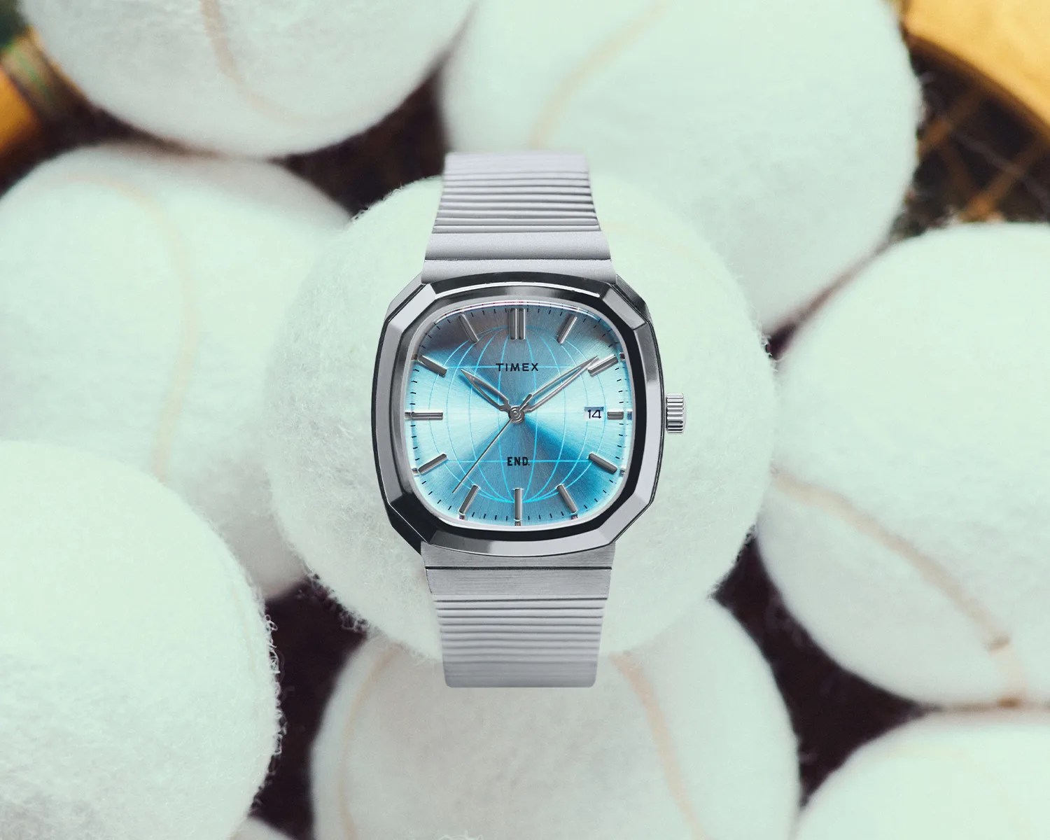 Silver Timex wristwatch with a blue globe-patterned dial and metal link band resting on white tennis balls.