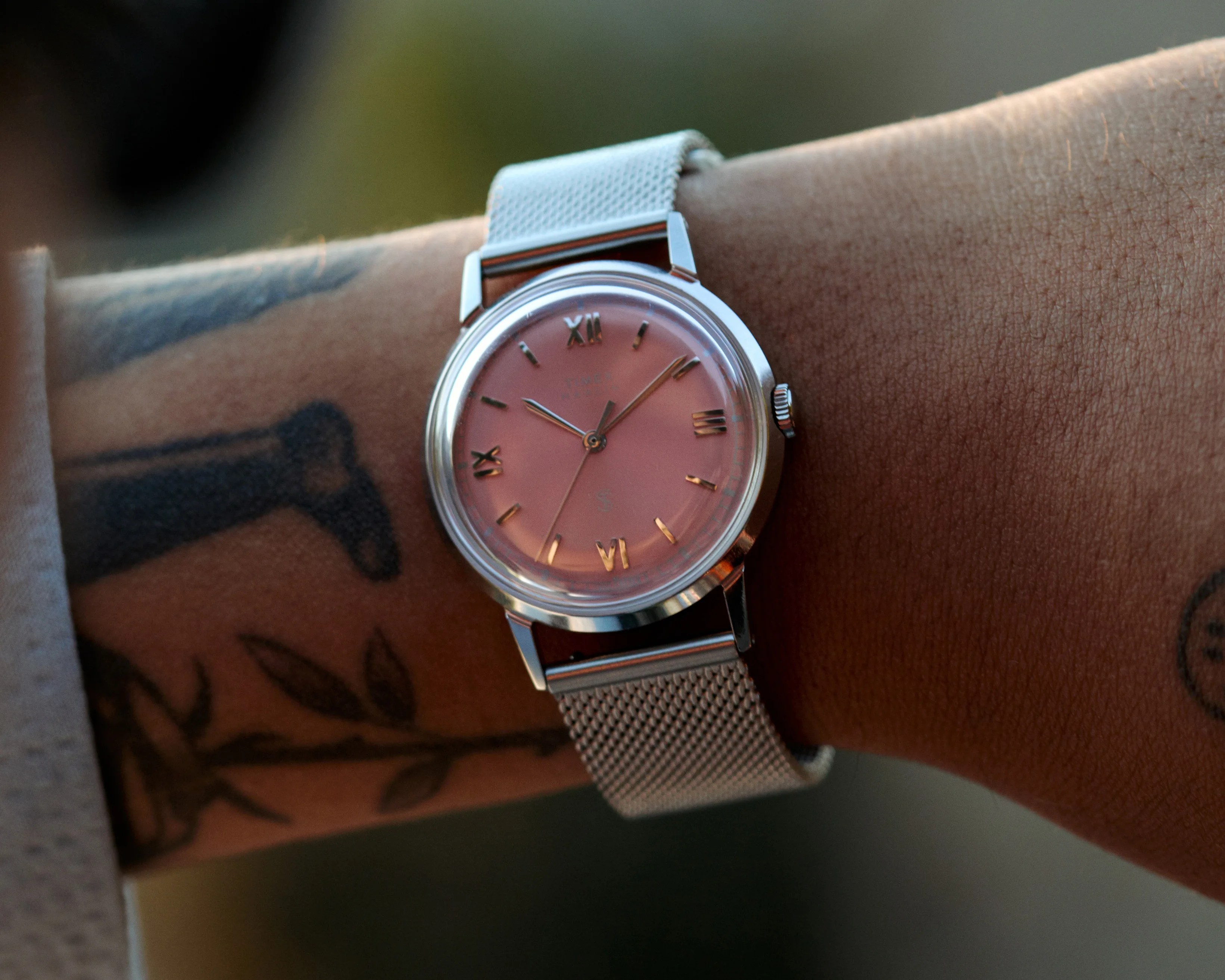Silver wristwatch with a pink dial and Roman numerals on a mesh metal strap worn on a tattooed wrist.